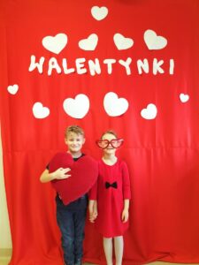 Zdjęcie przedstawia dzieci, które pozują do zdjęcia na ściance z napisem Walentynki. Dzieci trzymają w ręku czerwoną poduszkę w kształcie serca.