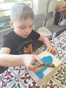 Zdjęcie przedstawia chłopca, który maluje na niebiesko swoją drewnianą szkatułkę.
