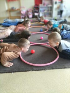 Zdjęcie przedstawia dzieci wykonujące ćwiczenie oddechowe, polegające na przesuwaniu piłeczki umieszczonej w obręczy za pomocą oddechu.