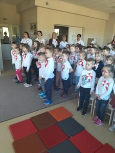 Zdjęcie przedstawia przedszkolaków zaproszonych na występ wraz z paniami, którzy stojąc na baczność śpiewają Hymn Polski.