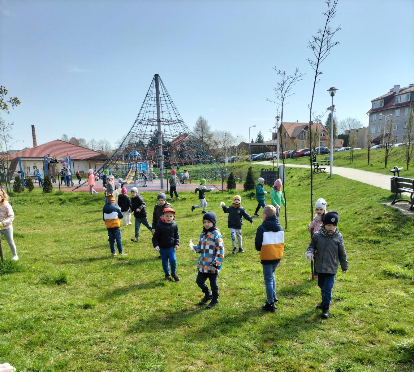 Zdjęcie przedstawia dzieci sprzątające teren przy pobliskim placu zabaw.