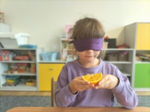 Zdjęcie przedstawia dziewczynkę, która za pomocą zmysłu smaku próbuje rozpoznać nazwę owocu. Dziewczynka ma zasłonięte oczy.