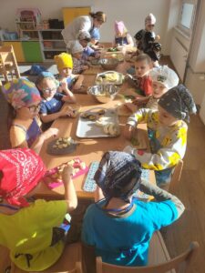 Zdjęcie przedstawia dzieci, które siedzą przy stole i kroją owoce do sałatki. Na głowie mają chustki, mają też ubrane fartuszki.