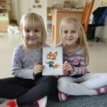Na zdjęciu widać dwie siedzące dziewczynki, które trzymają wspólnie kartkę obrazkową - nowego bohatera na zajęciach języka angielskiego Chester the fox, tzn. Lisa 🦊