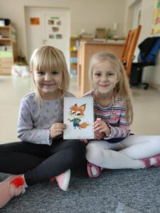 Na zdjęciu widać dwie siedzące dziewczynki, które trzymają wspólnie kartkę obrazkową - nowego bohatera na zajęciach języka angielskiego Chester the fox, tzn. Lisa 🦊