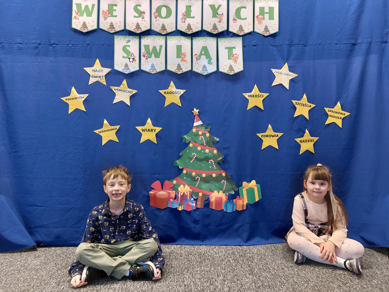 Chłopiec i dziewczynka pozują do zdjęcia, siedzą na dywanie. Za nimi znajduje się materiał, na którym wisi dekoracja i napis Wesołych Świąt.