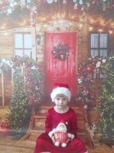 Na tle świątecznej chatki siedzi dziewczynka w mikołajkowej czapce i  trzyma małego mikołajka.  