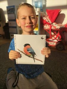 Na zdjęciu chłopiec prezentujący ilustrację z ptakiem przylatującym na zimę do Polski -gilem.
