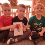Na zdjęciu można zauważyć trzech chłopców siedzących na dywanie w sali przedszkolnej. Chłopcy wspólnie trzymają kartę obrazkowa , tzw. flashcard, która ukazuje klauna - a clown.