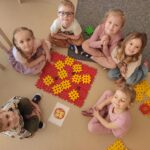 Zdjęcie ukazuje grupę dzieci siedzących na dywanie w małym kole. Po środku koła jest zbudowana pizza z klocków 'gofrow' . Zadaniem dzieci było zbudowanie pizzy takiej jaką jest widoczna za karcie flashcard - obrazującej pizzę.