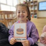 Zdjęcie przedstawia dziewczynkę prezentująca kartę obrazkową z tortem (birthday cake).