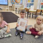 Trójka dzieci z karta obrazkową z jedną z zasad obowiązujących na zajęciach języka angielskiego. Dzieci ukazują tą zasadę. (Hands are still).