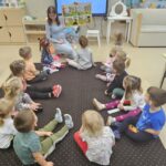 Zdjecoe ukazuje panią i dzieci siedzące w kole na dywanie. Pani trzyma w ręku duży plakat z histeoultjka obrazkową. Dziexi patrza na plakat o słuchają aktywnie historyjki.