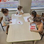 Zdjecia ukazuje dzieci siedzące przy stoliku. Malują one numer alarmowy czerwoną farbą.