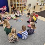 Zdjęcie ukazuje dzieci siedzących w kólku na dywanie. Dzieci słuchają opowiadania o kredkach.