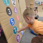 Dziecko uczestniczy w zabawie ruchowej 'Windzie'. Na zdjęciu chłopiec wsjazuje liczbę 7. 