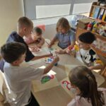 Dzieci przy stoliku malują farbami papugę.