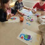 Dzieci przy stoliku malują farbami papugę.