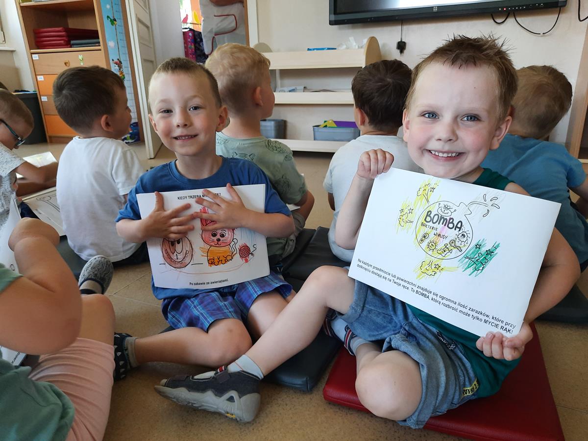 Dwoje chłopów prezentuje swoją pracę, pokolorowane ilustracje wirusów w kolorowance edukacyjnej. Za chłopcami siedzą inne dzieci.