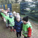 Zdjęcie przedstawia grupę dzieci3-4 letnich na spacerze. Dzieci stoją ustawione w parach, mają także kamizelki odblaskowe.