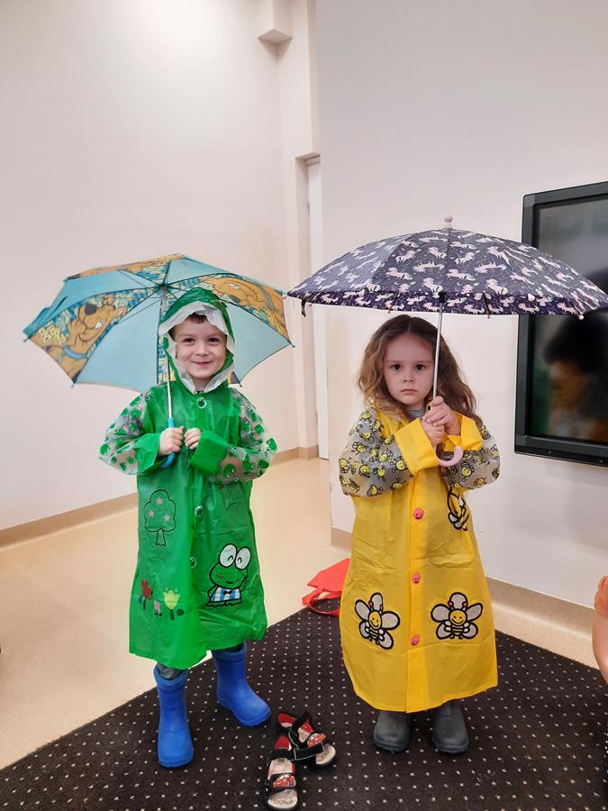 Chłopiec i dziewczynka demonstrują ubiór odzieży na deszczową pogodę. Mają ubrane: deszczaki i kalosze, w ręku trzymają rozłożoną parasolkę.