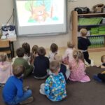 Zdjęcie ukazuję grupę dzieci, siedzących w sali przedszkolnej. Dzieci oglądają wersję wizualną piosenki pt.: 'Der Spielzeug Tanz' na tablicy interaktywnej.