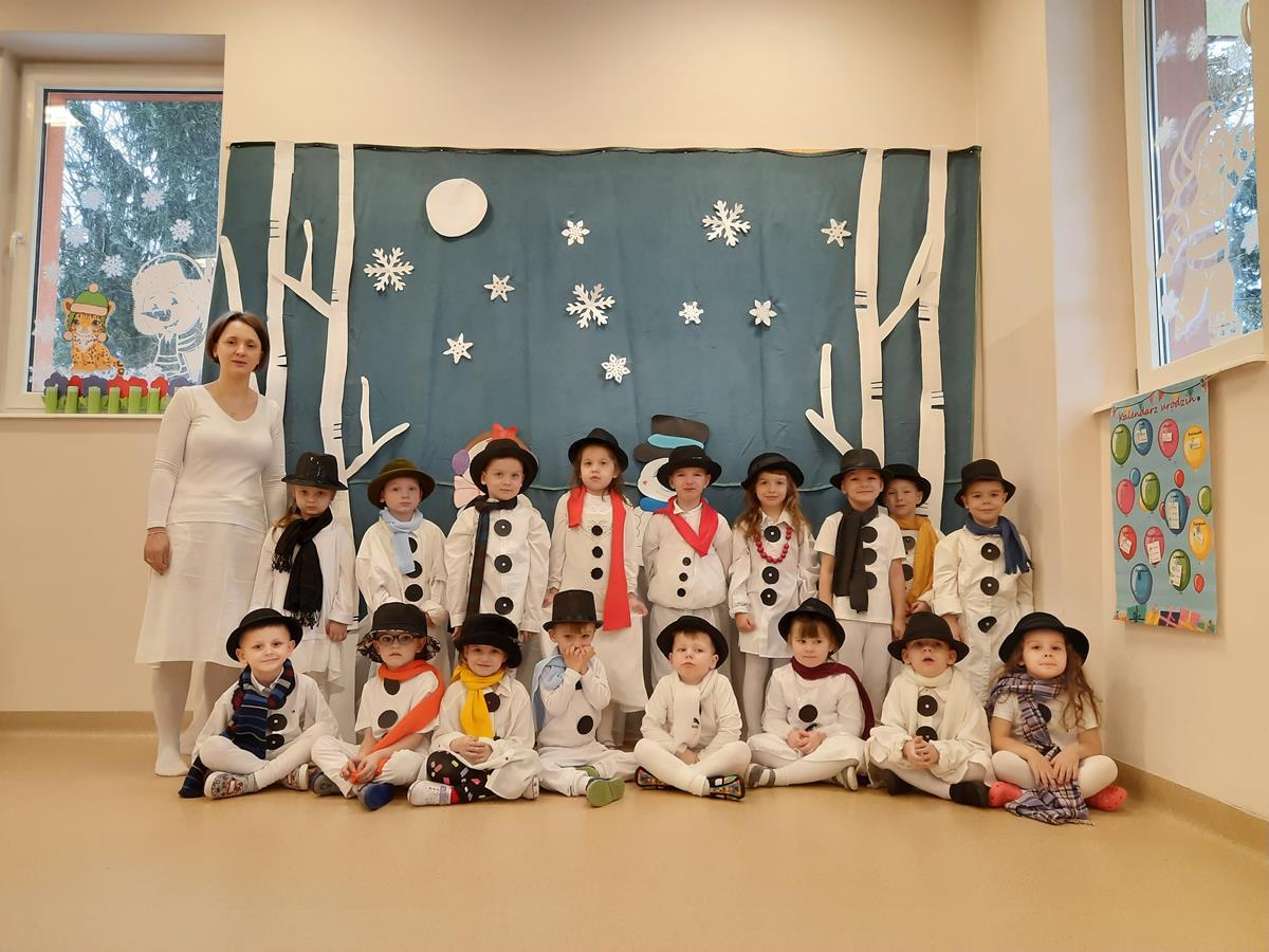 Dzieci wraz z nauczycielem pozują do grupowego zdjęcia. Dzieci przebrane są za bałwanki: mają białe spodnie i bluzi, czarne kropki na bluzce, czarny kapelusz na głowie i kolorowy szal na szyi. W tle wisi zimowa dekoracja.