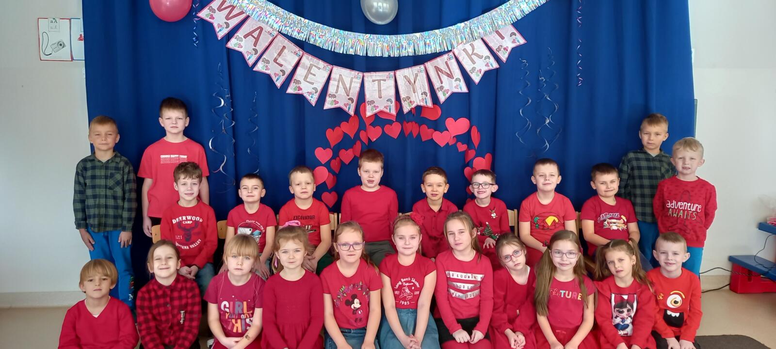 Grupa Delfinki pozuje do zdjęcia ubrana na kolor czerwony, w tle dzieci napis "Walentynki" oraz dekoracja serduszek.