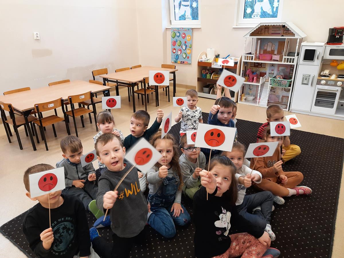 Dzieci siedzą na dywanie. W rękach trzymają tabliczki ze smutną czerwona minką. Jest to odpowiedź na zadawane pytanie.