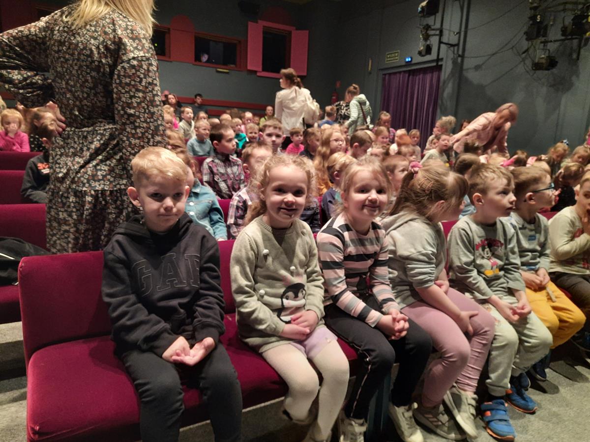 5 dzieci z grupy Panterki siedzą na kanapach czekając na spektakl. W tle siedzą pozostali goście teatru.