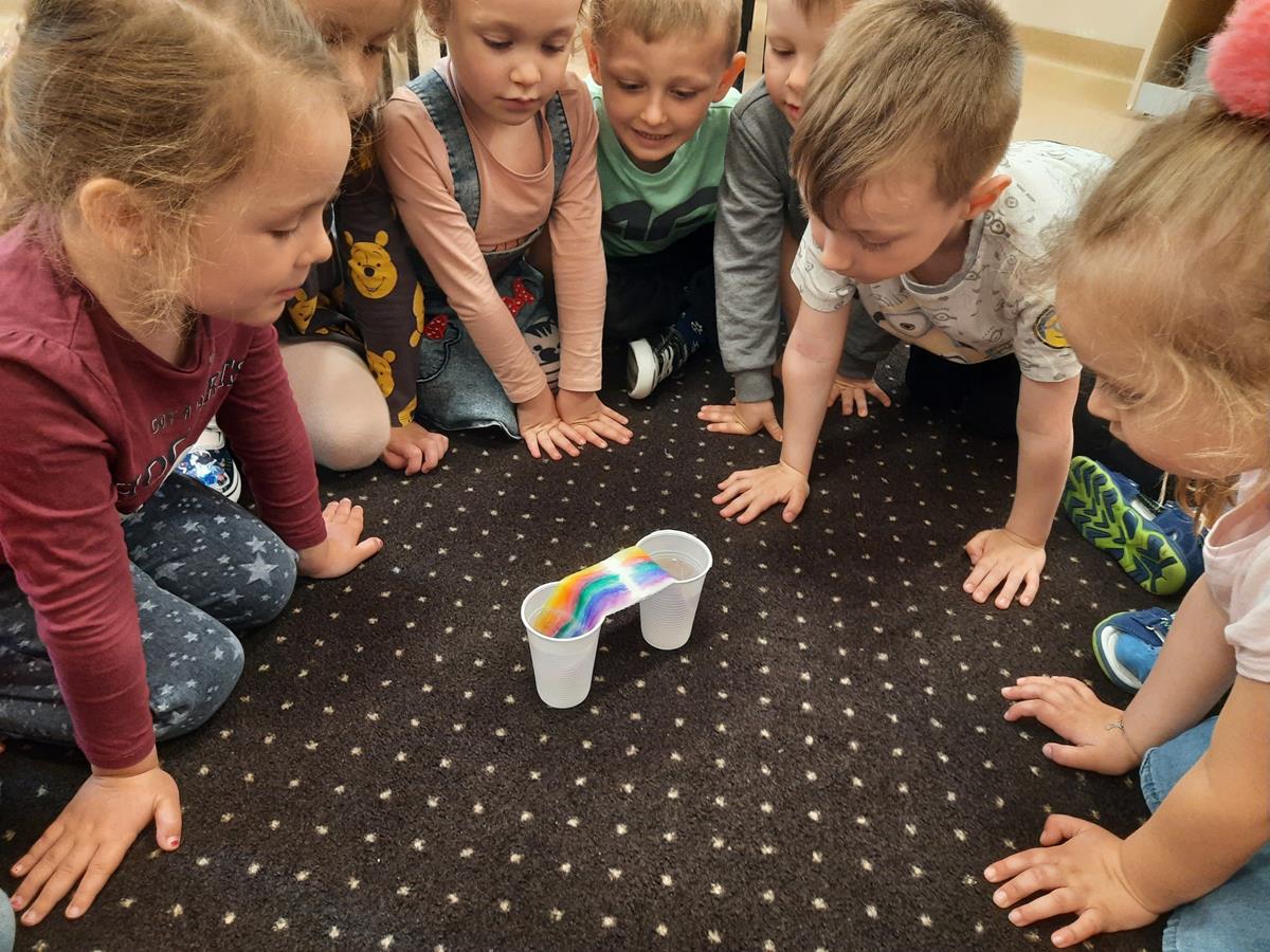 Dzieci siedzą w kole na dywanie. W środku w dwóch kubkach są kolorowe papiery. Dzieci wykonują eksperyment kolorowa tęcza.