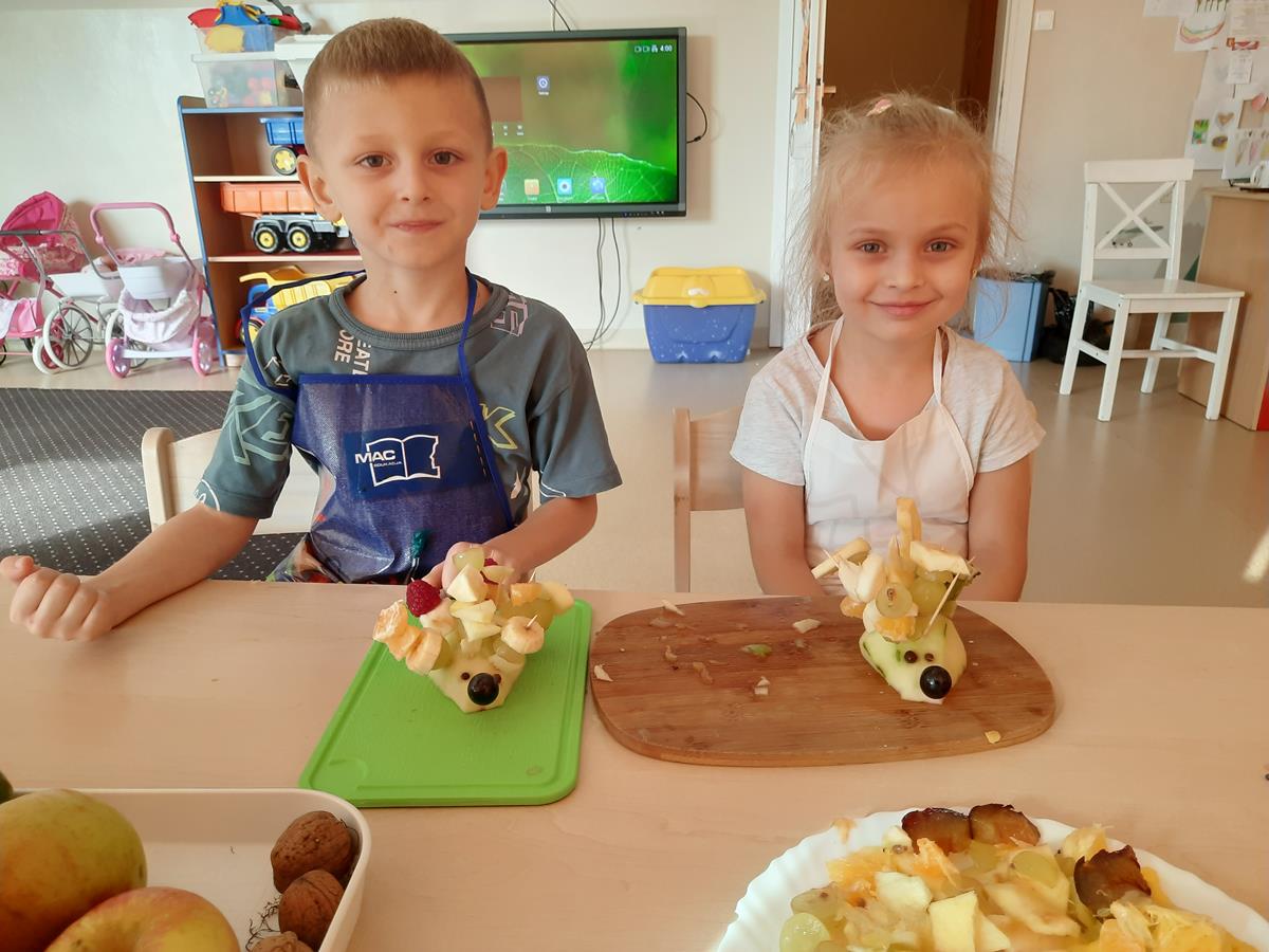 Chłopiec i dziewczynka siedzą przy stoliku. Prezentują wykonane przez siebie jeżyki z owoców.