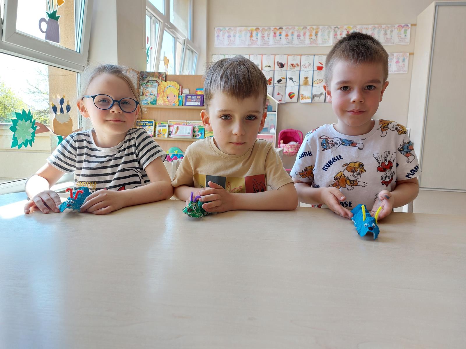 Troje dzieci siedzi przy stoliku i prezentują wykonane przez siebie gąsienice.