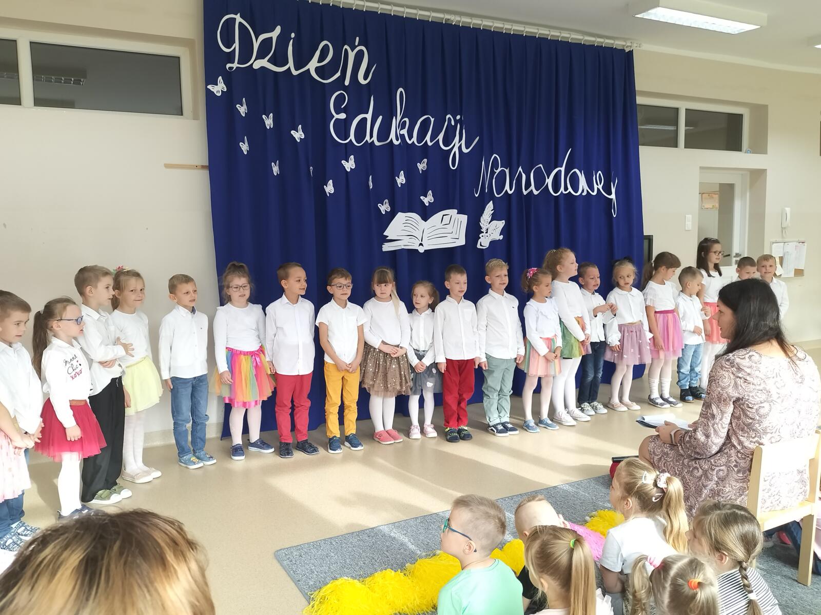 Zdjęcie przedstawia dzieci podczas występu. Dzieci ustawione są w półkolu. Ubrane są w białe bluzeczki. Nad nimi widnieje dekoracja "Dzień Edukacji Narodowej".