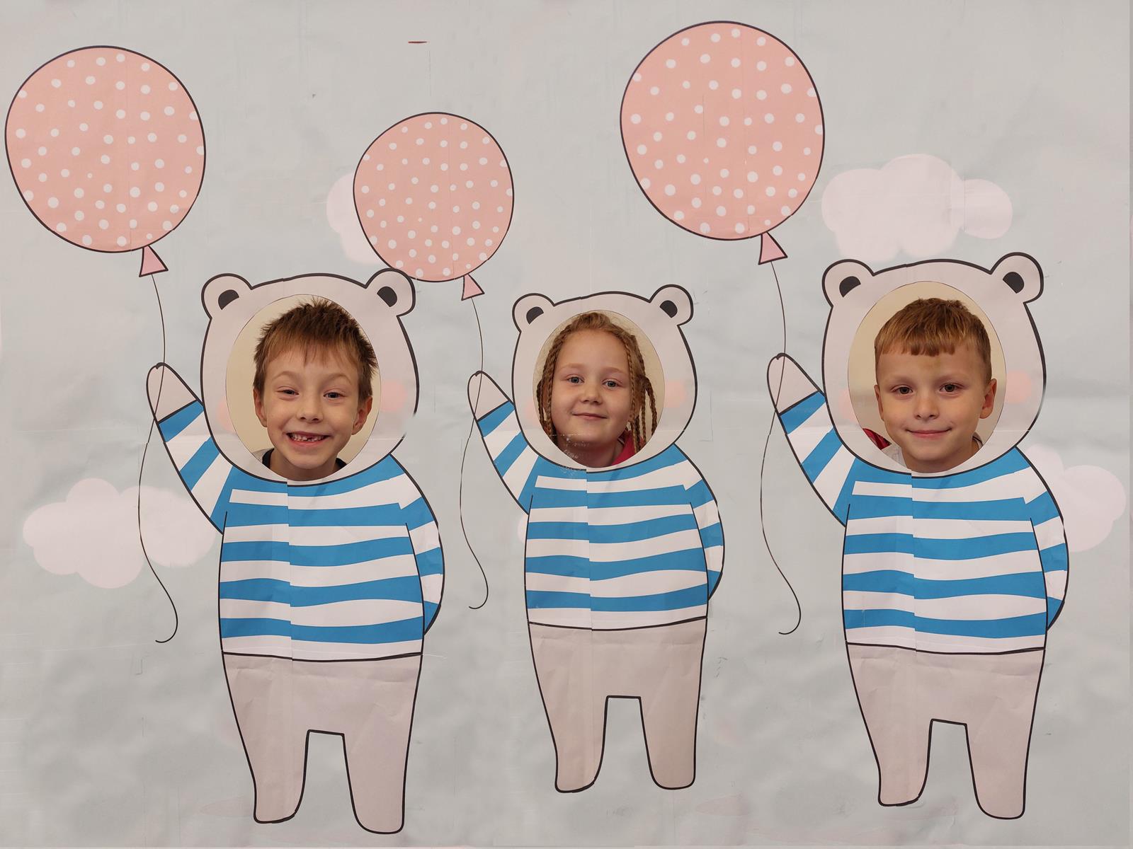 Troje dzieci pozuje w fotobudce misiów, na dużym plakacie są narysowane misie, dzieci patrzą przez dziurę, widać tylko ich głowy.