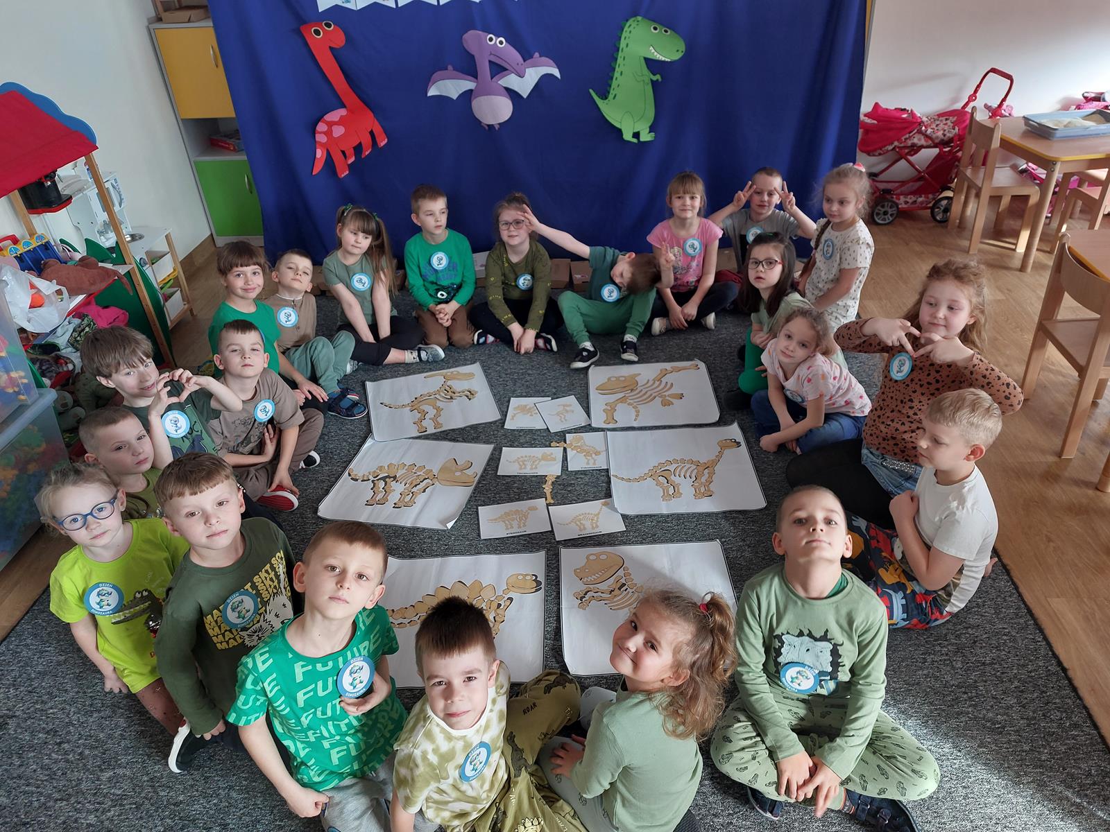 Dzieci siedzą w kole na dywanie, między nimi leżą ułożone szkielety dinozaurów.