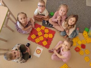 Zdjęcie ukazuje grupę dzieci siedzących na dywanie w małym kole. Po środku koła jest zbudowana pizza z klocków 'gofrow' . Zadaniem dzieci było zbudowanie pizzy takiej jaką jest widoczna za karcie flashcard - obrazującej pizzę.