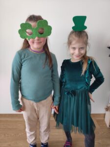 Zdjęcie ukazuje dwie osoby stojące - dziewczynki mają ubrane na sobie rekwizyty związane ze Dniem Świętego Patryka : a top hat and a shamrock glasses.