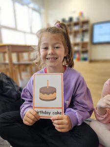 Zdjęcie przedstawia dziewczynkę prezentująca kartę obrazkową z tortem (birthday cake).