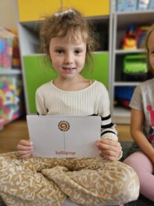 Na zdjęciu widzimy dziewczynkę prezentująca kartę obrazkową z lizakiem (lollipops).