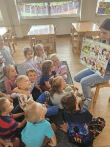 Dzieci siedzą na dywanie, patrzą na plakat i słuchają historyjki.