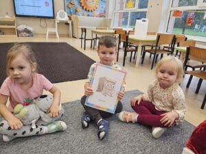 Trójka dzieci z karta obrazkową z jedną z zasad obowiązujących na zajęciach języka angielskiego. Dzieci ukazują tą zasadę. (Hands are still).