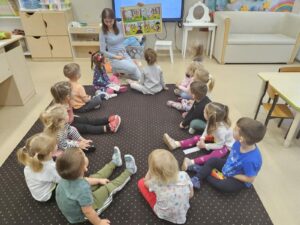 Zdjecoe ukazuje panią i dzieci siedzące w kole na dywanie. Pani trzyma w ręku duży plakat z histeoultjka obrazkową. Dziexi patrza na plakat o słuchają aktywnie historyjki.