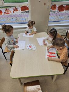 Zdjecia ukazuje dzieci siedzące przy stoliku. Malują one numer alarmowy czerwoną farbą.