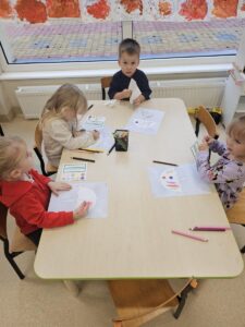 Dzieci siedzą przy stoliku, kolorują kartę pracy, na której jest parasolka.