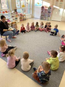 Dzieci siedzą w kole wraz z panią. Uważnie jej słuchają.
