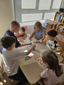 Dzieci przy stoliku malują farbami papugę.