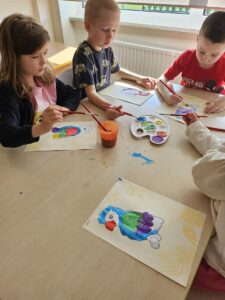 Dzieci przy stoliku malują farbami papugę.