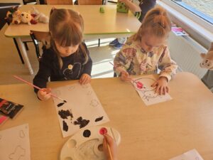 Dwójka dzieci maluje farbami przy stoliku. Jedna dziewczynka łaty krowy , a druga głowę.